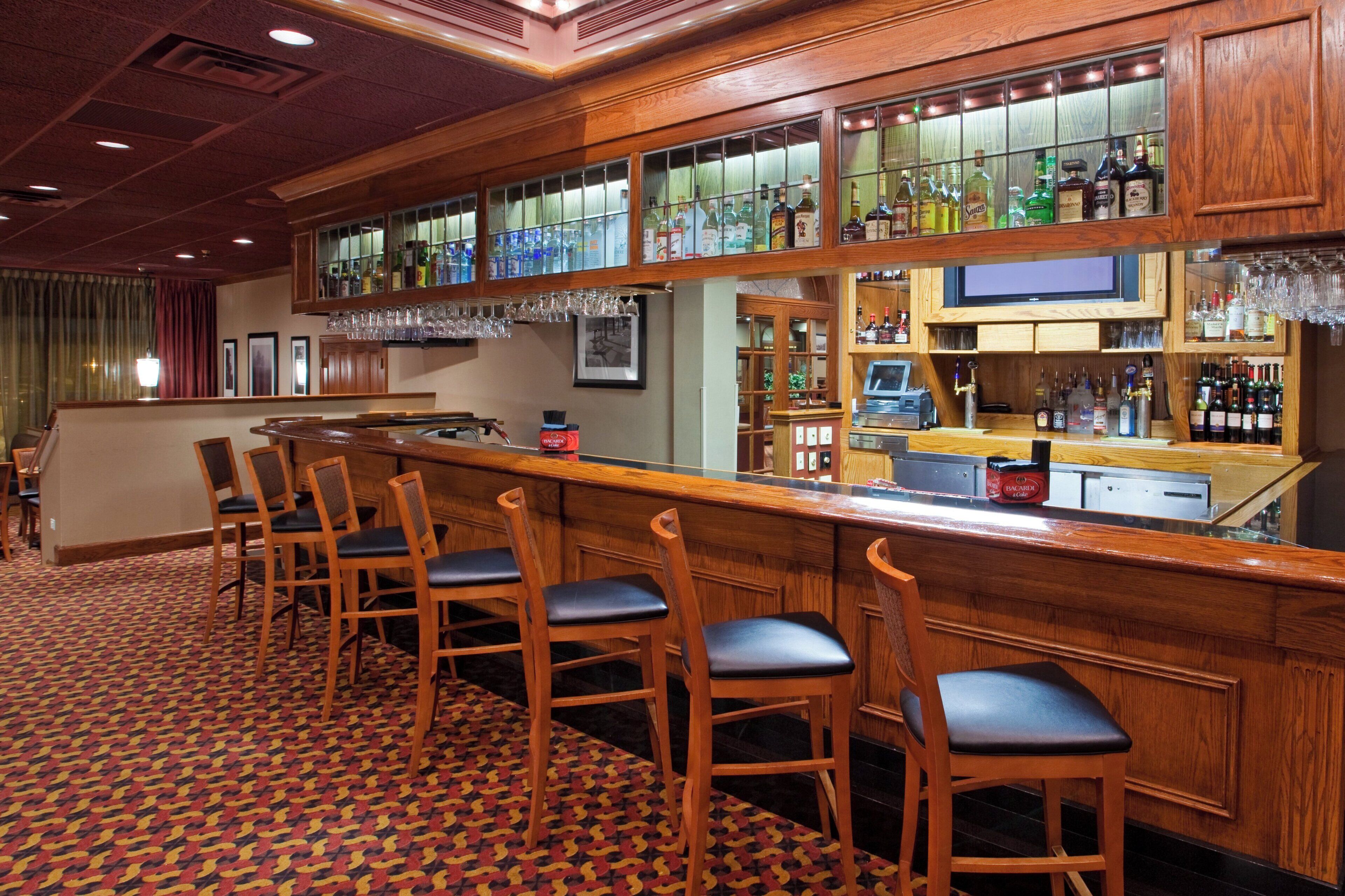 Uma área de bar com banquetas de madeira, um balcão com um registradora e prateleiras cheias de garrafas.