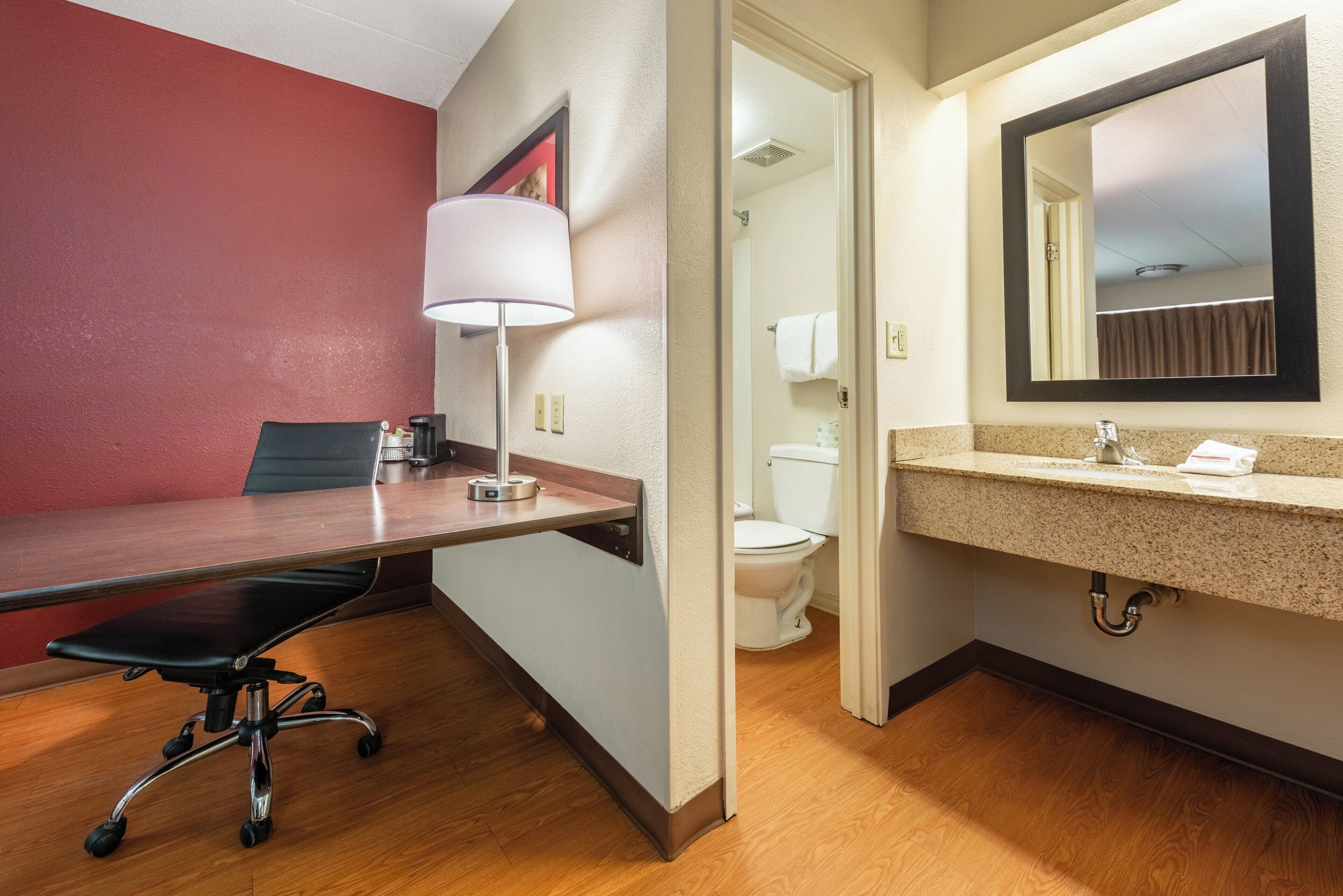 A hotel room with a wooden desk, a chair, a lamp, a toilet, and a sink with a mirror.