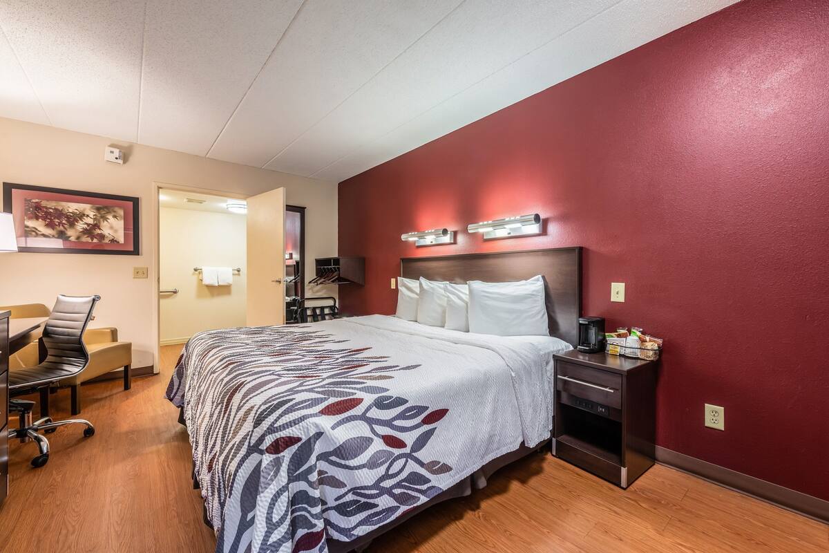 A hotel room with a bed, a desk with a chair, a nightstand, and a wall-mounted light fixture.