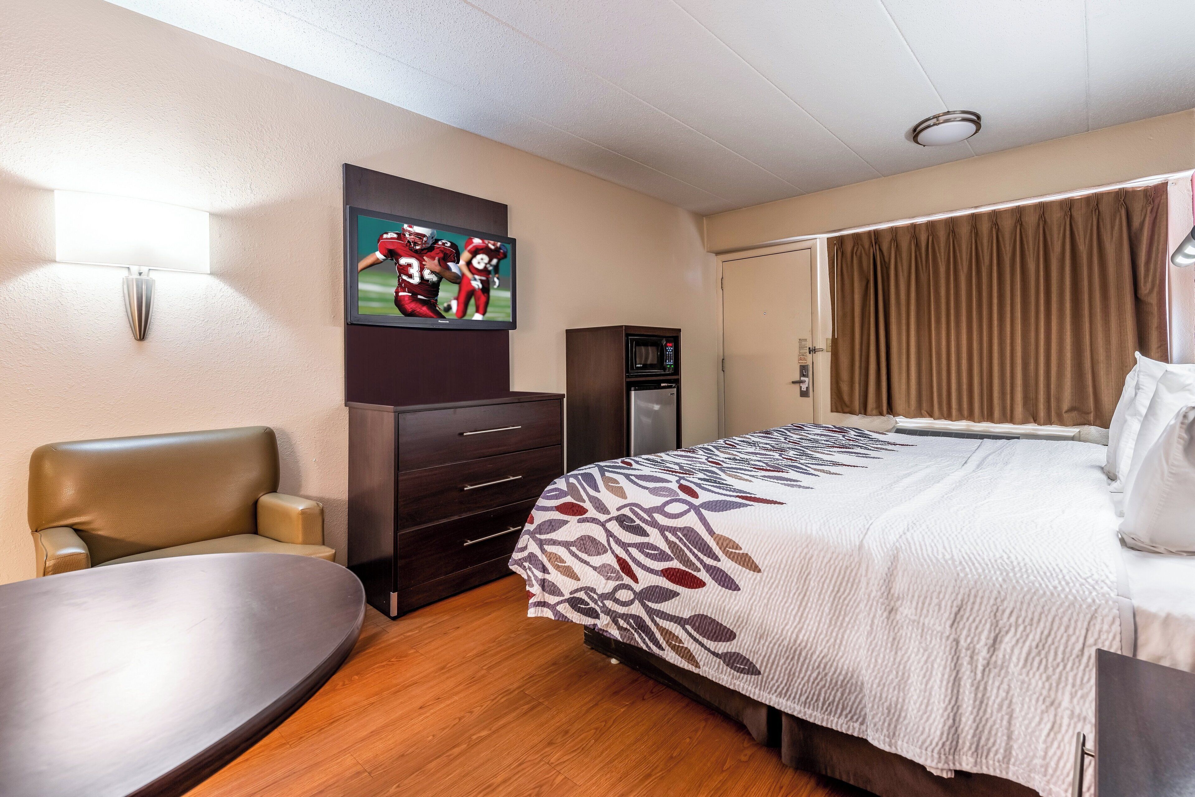 Habitación de hotel con cama, escritorio, silla, televisor montado en la pared, microondas y puerta con cortinas.