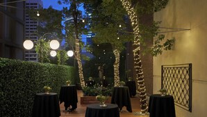 Un área de comedor al aire libre con mesas cubiertas con manteles negros, rodeada de follaje e iluminación con luces colgantes.