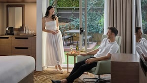 Habitación de hotel moderna con cama, zona de estar con mesa, ventana grande con cortinas y vistas a zonas verdes.