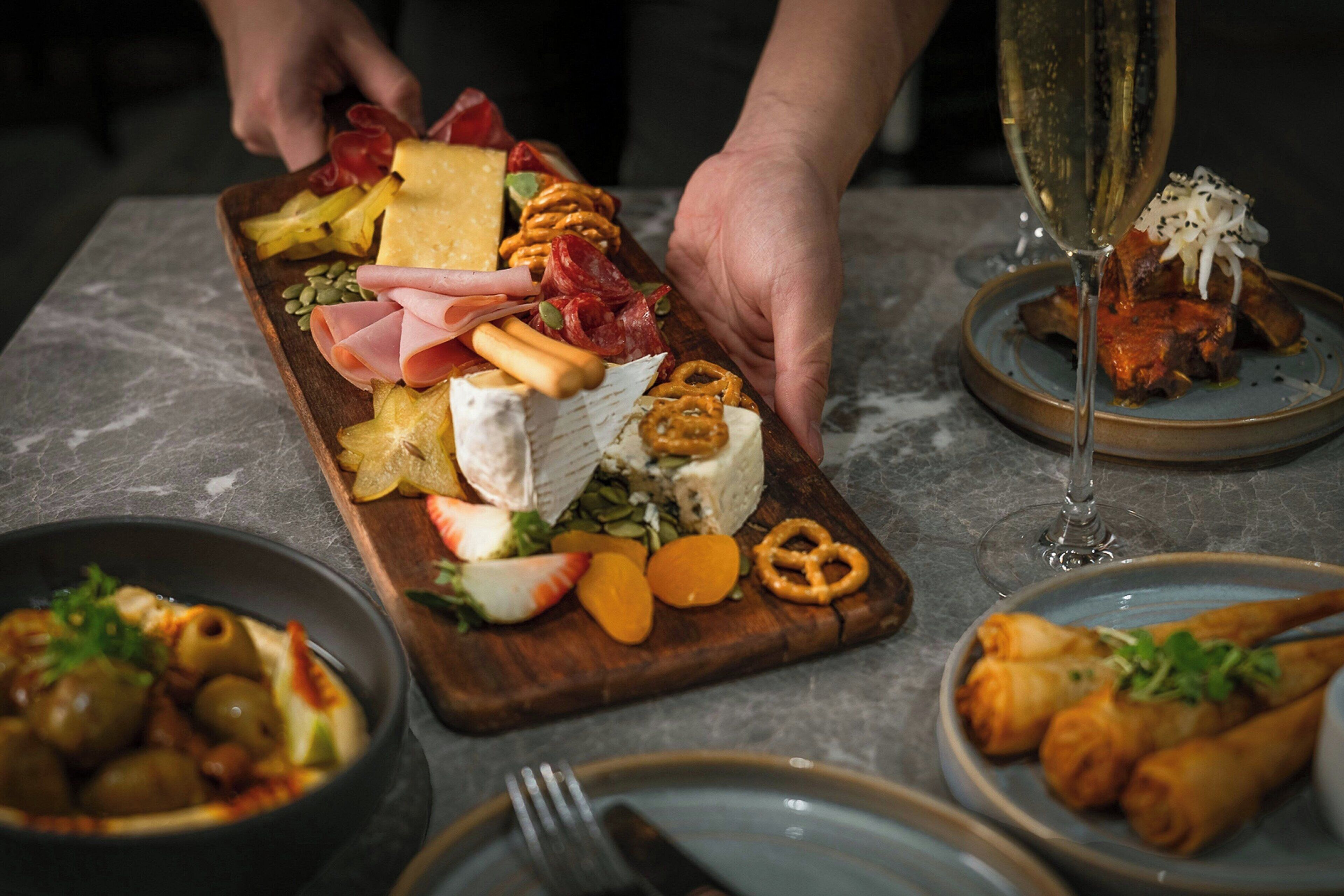 A wooden board with various cheeses, meats, and vegetables, a glass of champagne, and a bowl of olives.
