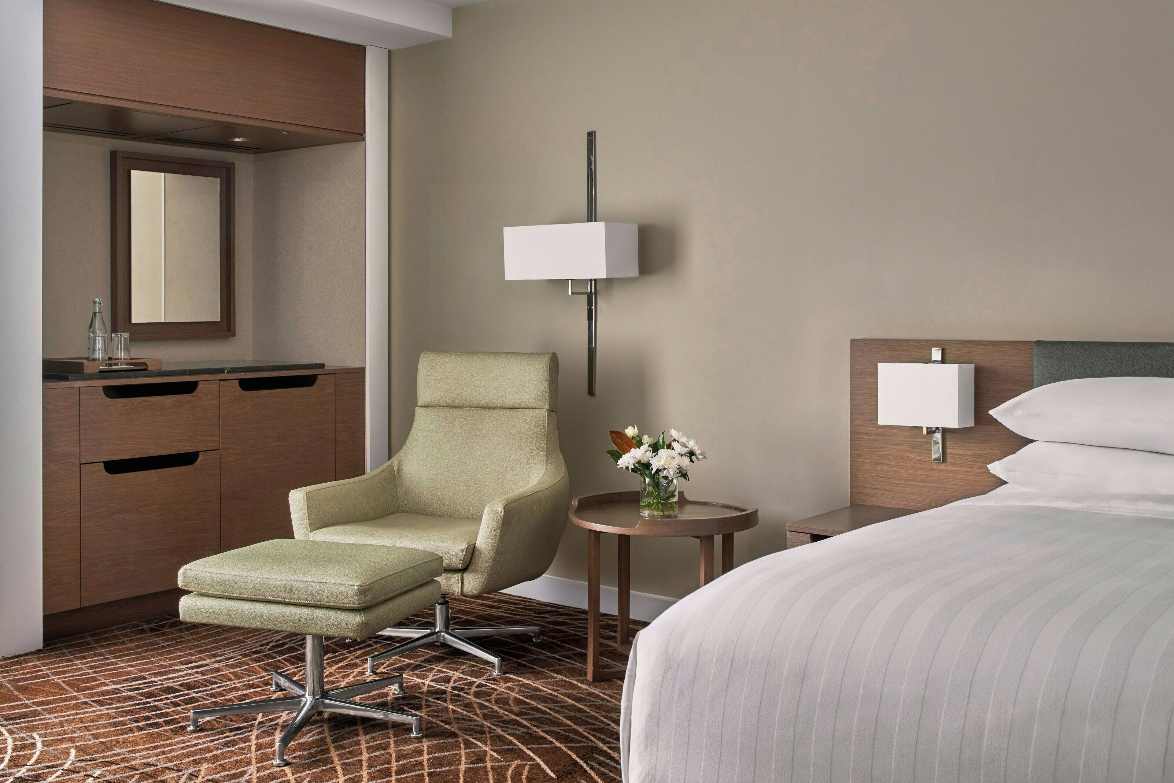 A hotel room with a bed, a desk with a lamp, a chair, a small table with a vase of flowers, and a mirror on the wall.