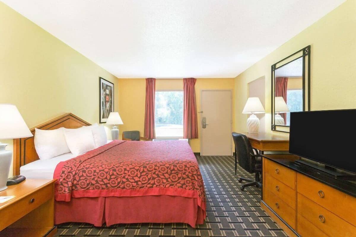 A hotel room with a bed, a desk with a TV, a chair, a desk lamp, a window with curtains, and a door.