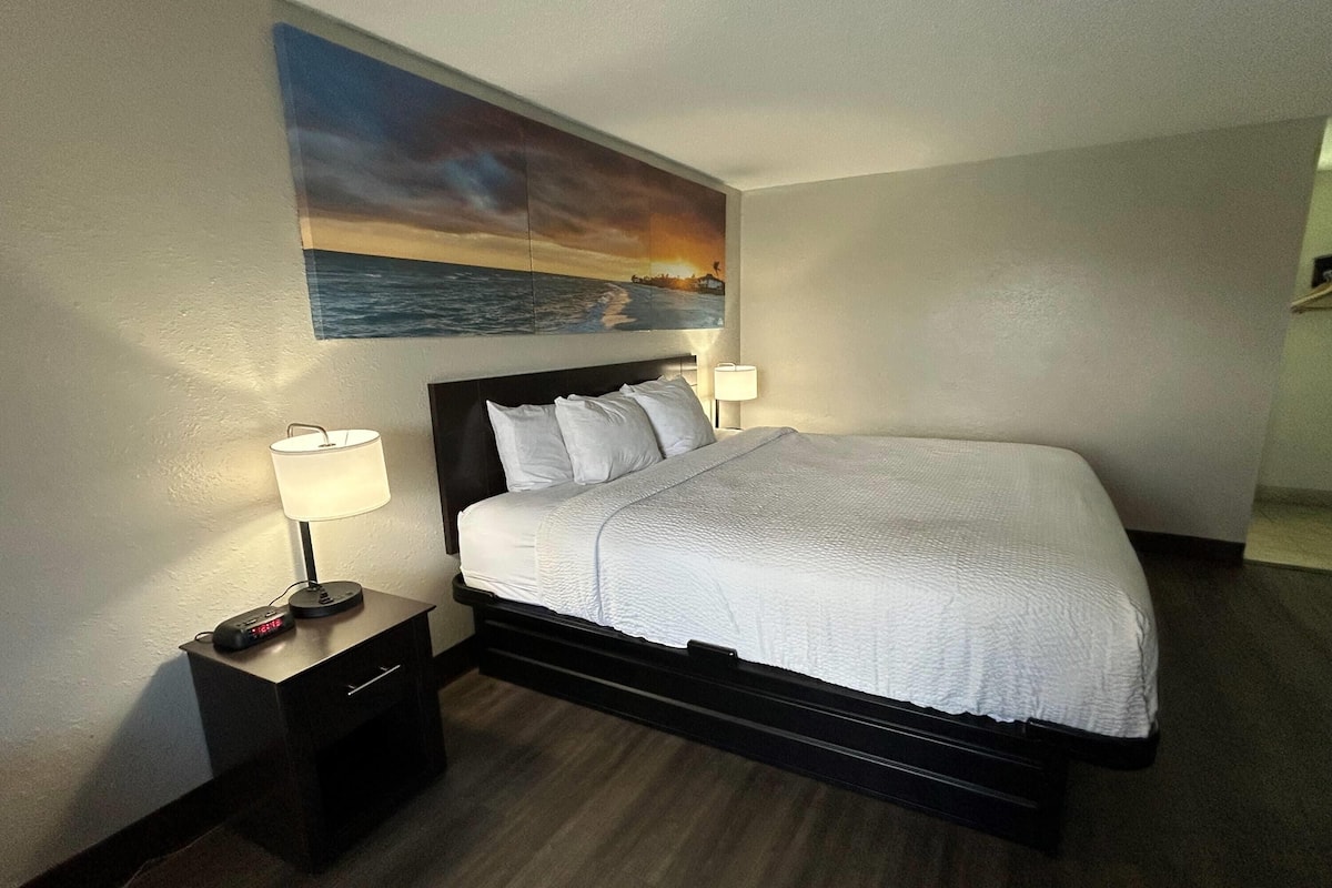 A hotel room with a bed, nightstands, a painting of a sunset over the ocean, and a lamp.