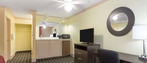 A hotel room with a ceiling fan, a desk with a chair, a TV, a microwave, and a small kitchenette with a sink and cabinets.