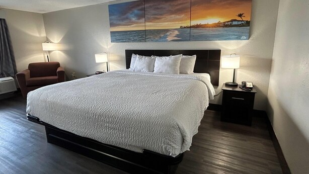 A hotel room with a large bed, two bedside tables with lamps, a chair, and a wall-mounted painting of a sunset over the ocean. - Days Inn by Wyndham Savannah Airport (Garden City)
