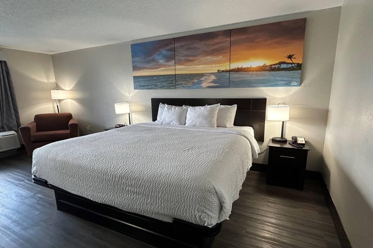 A hotel room with a large bed, two bedside tables with lamps, a chair, and a wall-mounted painting of a sunset over the ocean.