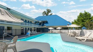 A swimming pool with lounge chairs and umbrellas, a building with a blue roof, and a fenced area with a white fence. - Days Inn by Wyndham Savannah Airport (Garden City)