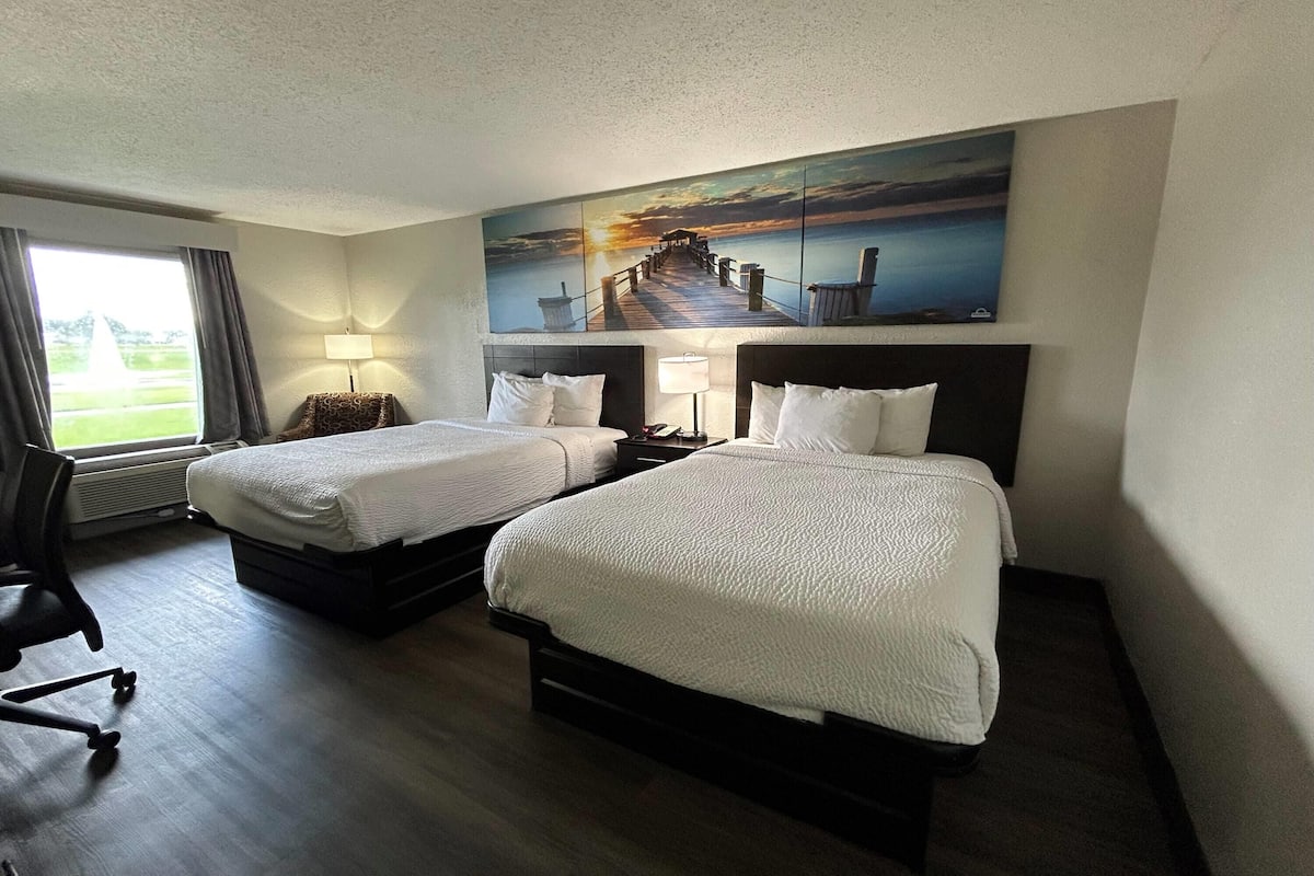 A hotel room with two beds, a desk, a chair, a lamp, and a large painting of a pier.