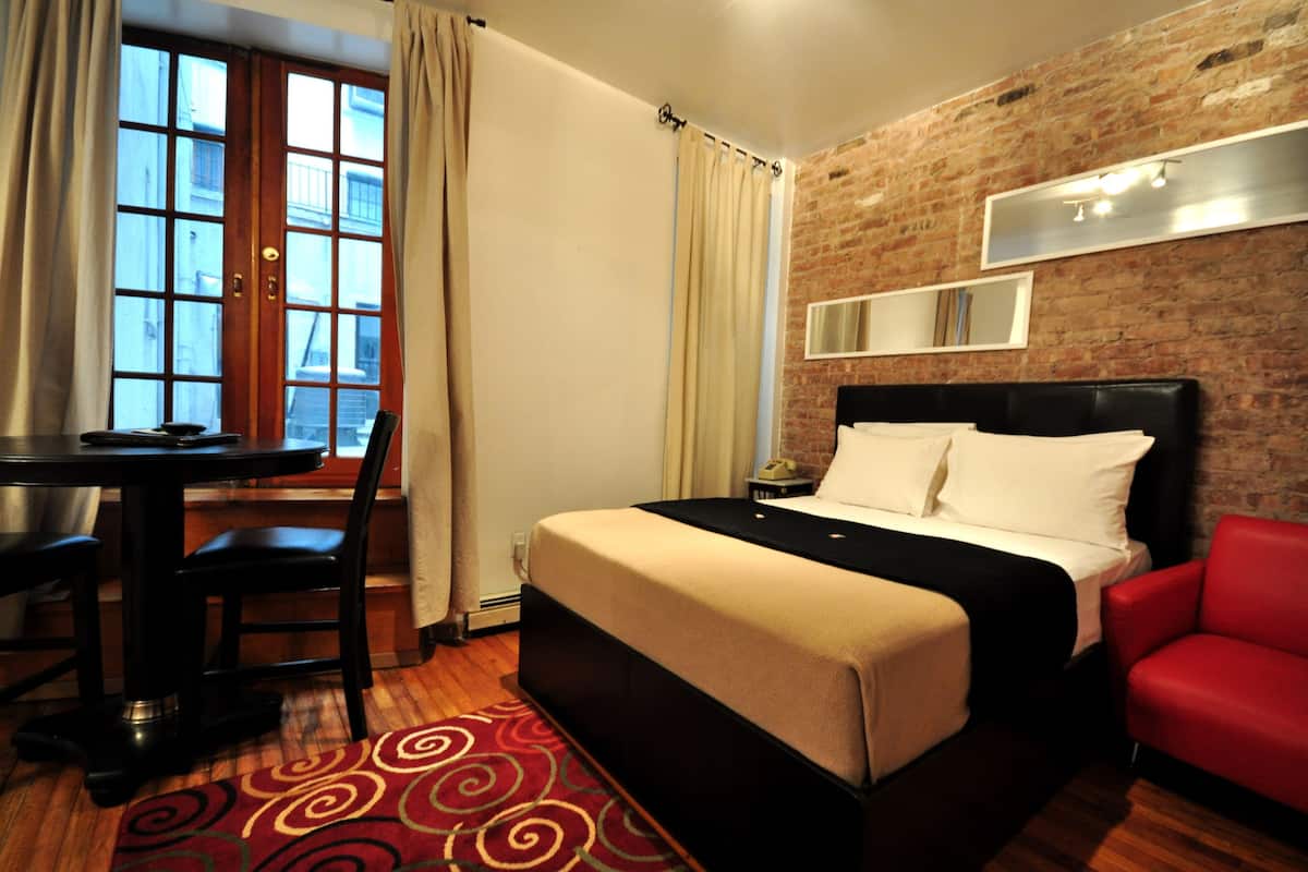 A hotel room with a bed, a desk, a chair, a red armchair, a brick wall, and a window with curtains.