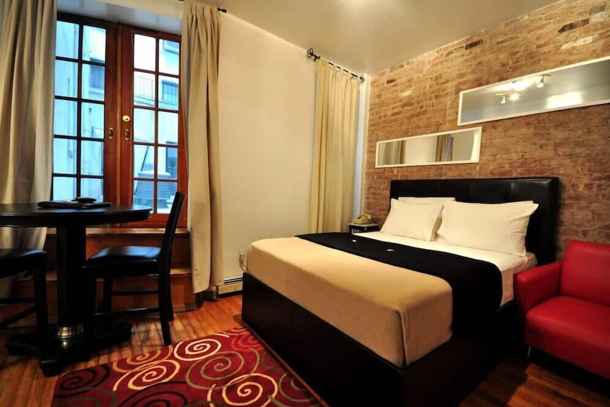 A hotel room with a bed, a desk, a chair, a red armchair, a brick wall, and a window with curtains.