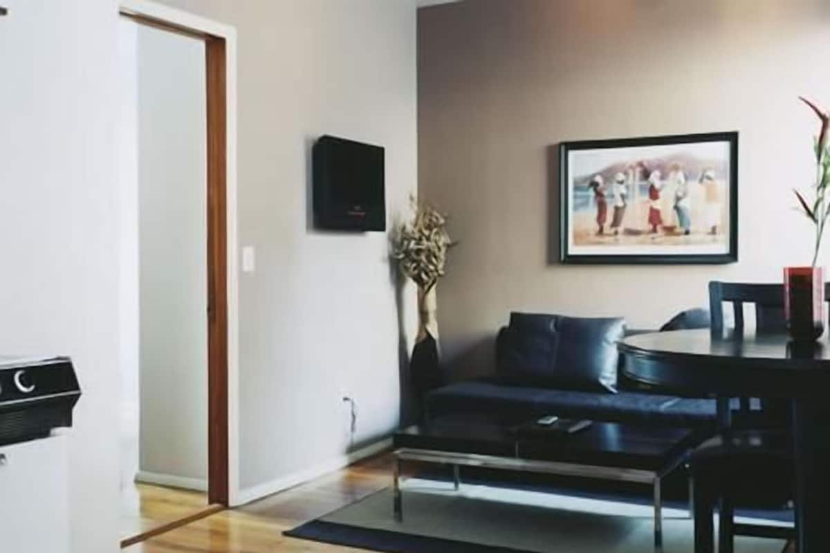 A living room with a dark-colored sofa, a coffee table, and a dining table with chairs. There is a framed picture on the wall and a small plant on the coffee table.