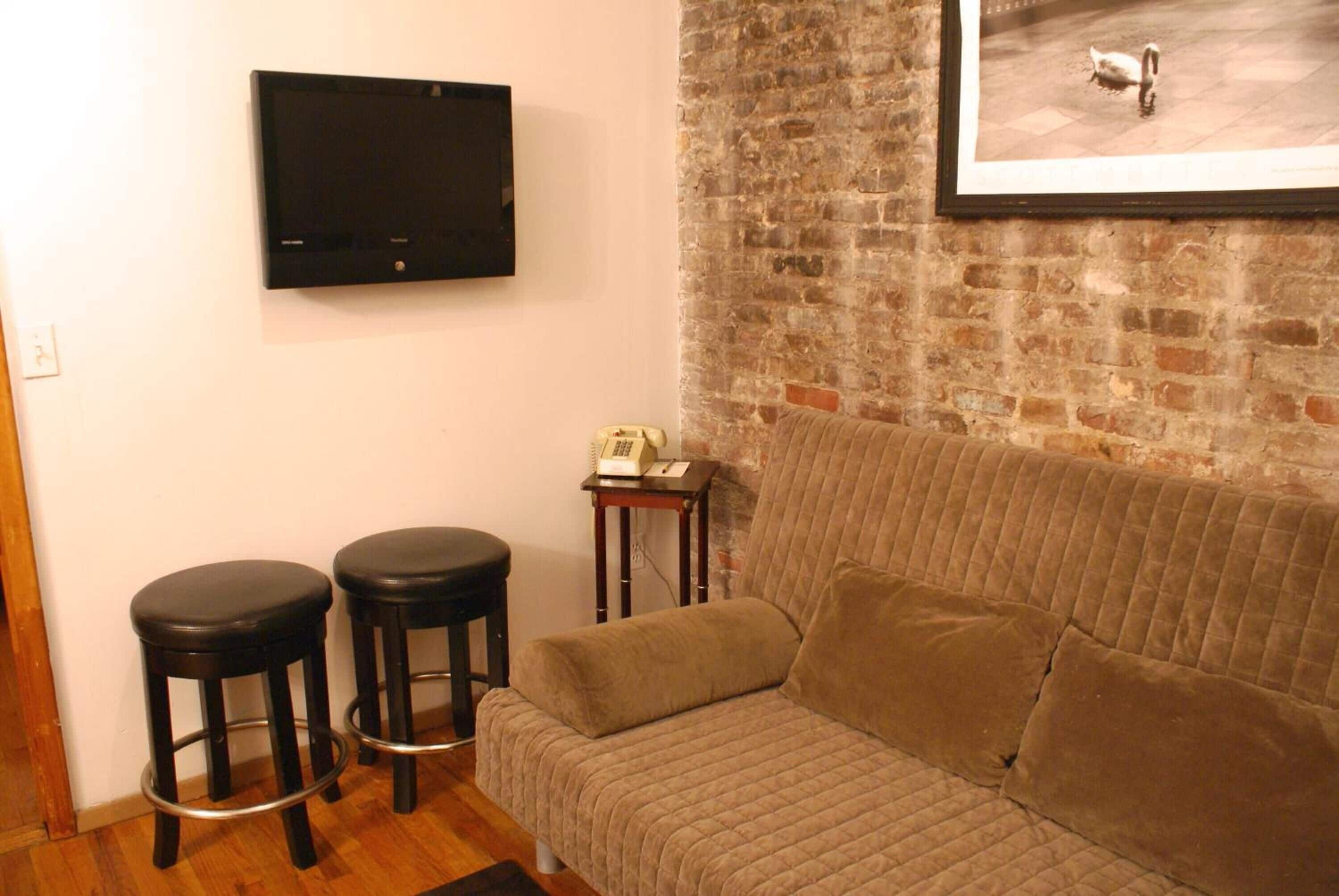 A living room with a brown sofa, two black bar stools, a flat-screen TV mounted on the wall, and a framed picture of a swan on the brick wall.