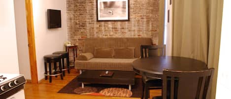 Una sala de estar acogedora con pared de ladrillo, un sofá, una mesa de centro, un televisor y un comedor con sillas y una mesa.