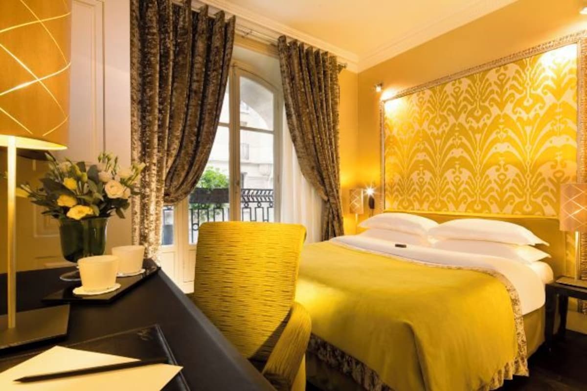 A hotel room with a bed, a desk with a chair, a vase with flowers, and a window with curtains.