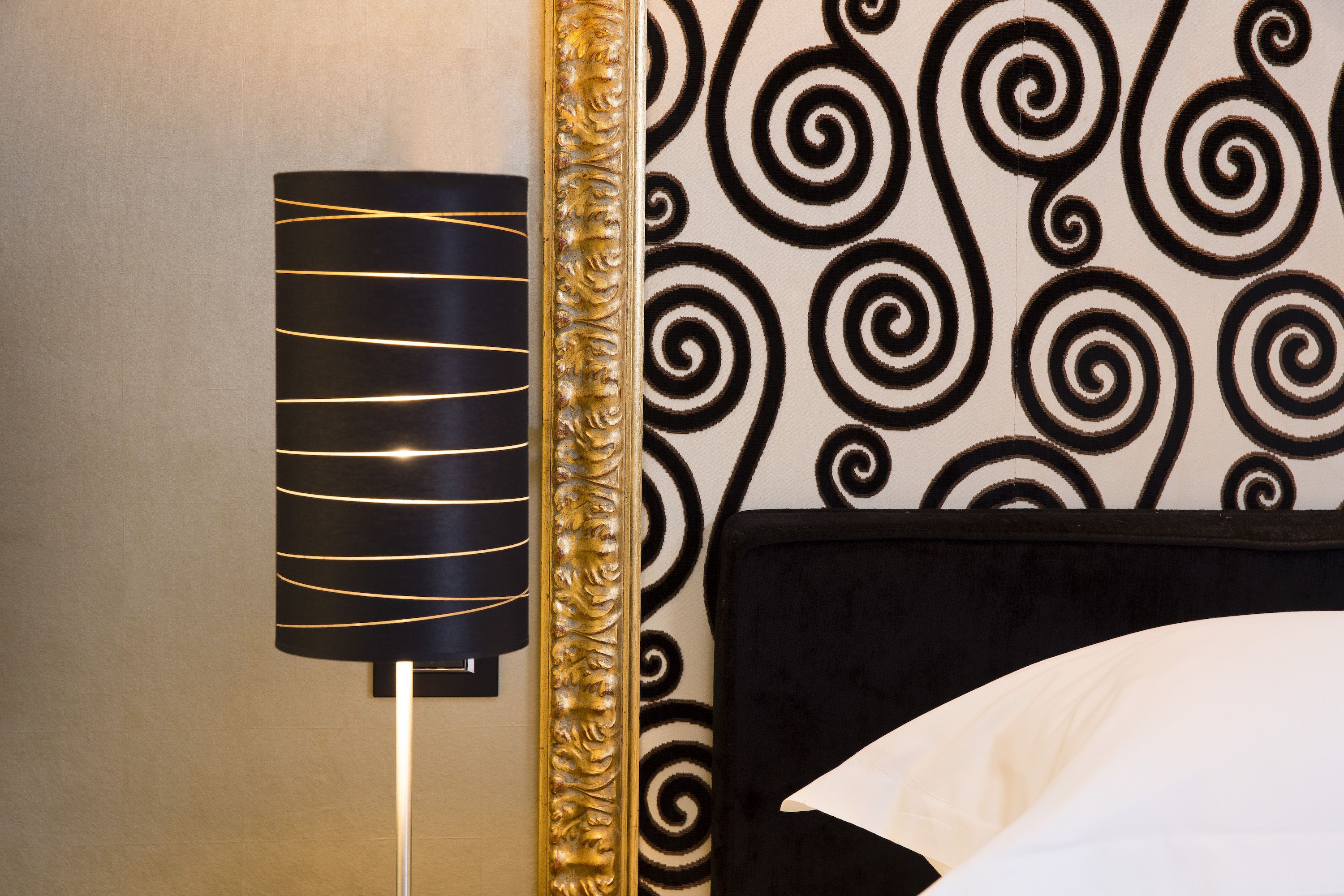 Um quarto com papel de parede estampado em preto e branco, uma luminária preta com detalhes dourados e um espelho com moldura dourada.