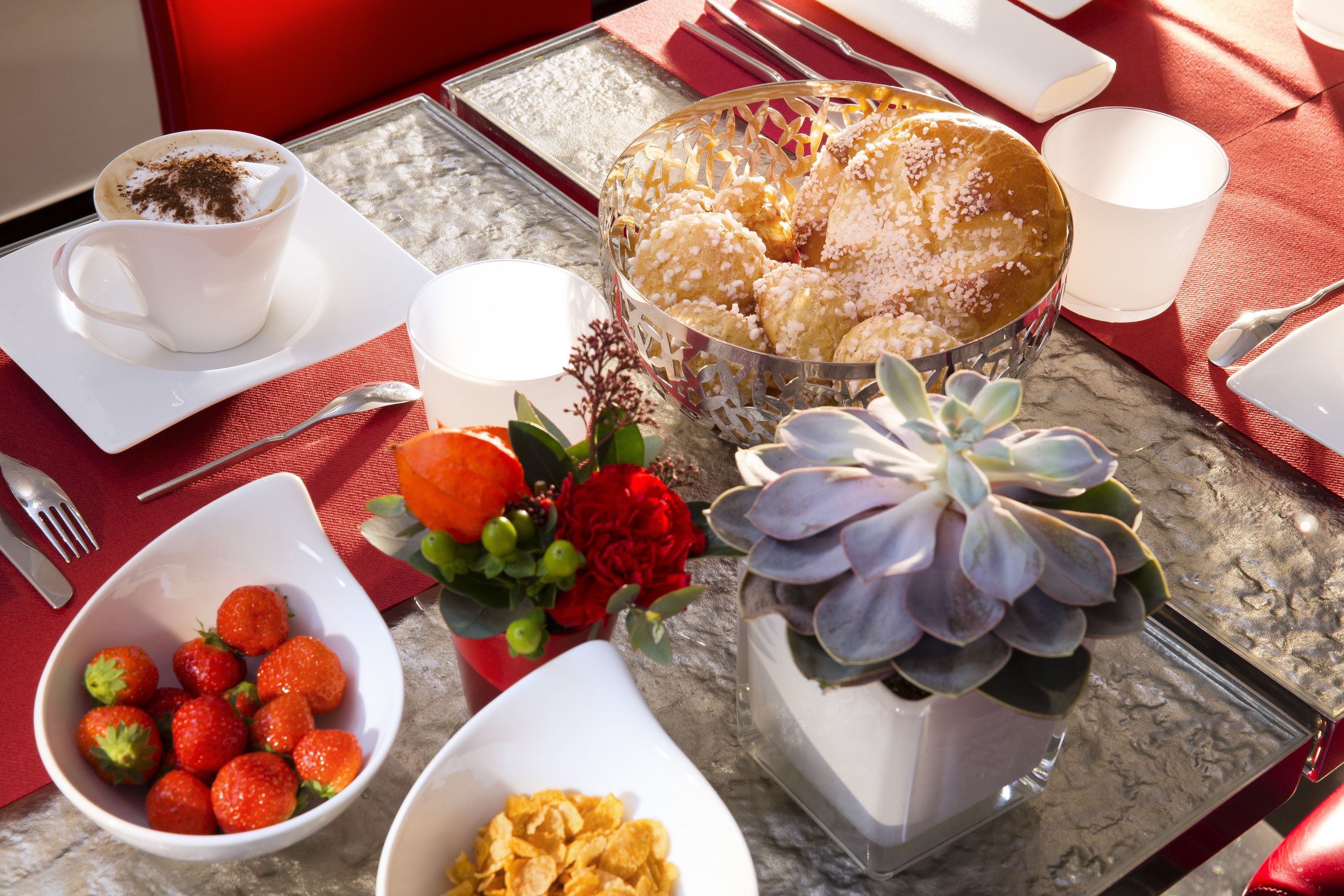 Une table de petit-déjeuner avec une tasse de café, un bol de fraises, un bol de céréales, un gâteau et une plante succulente.