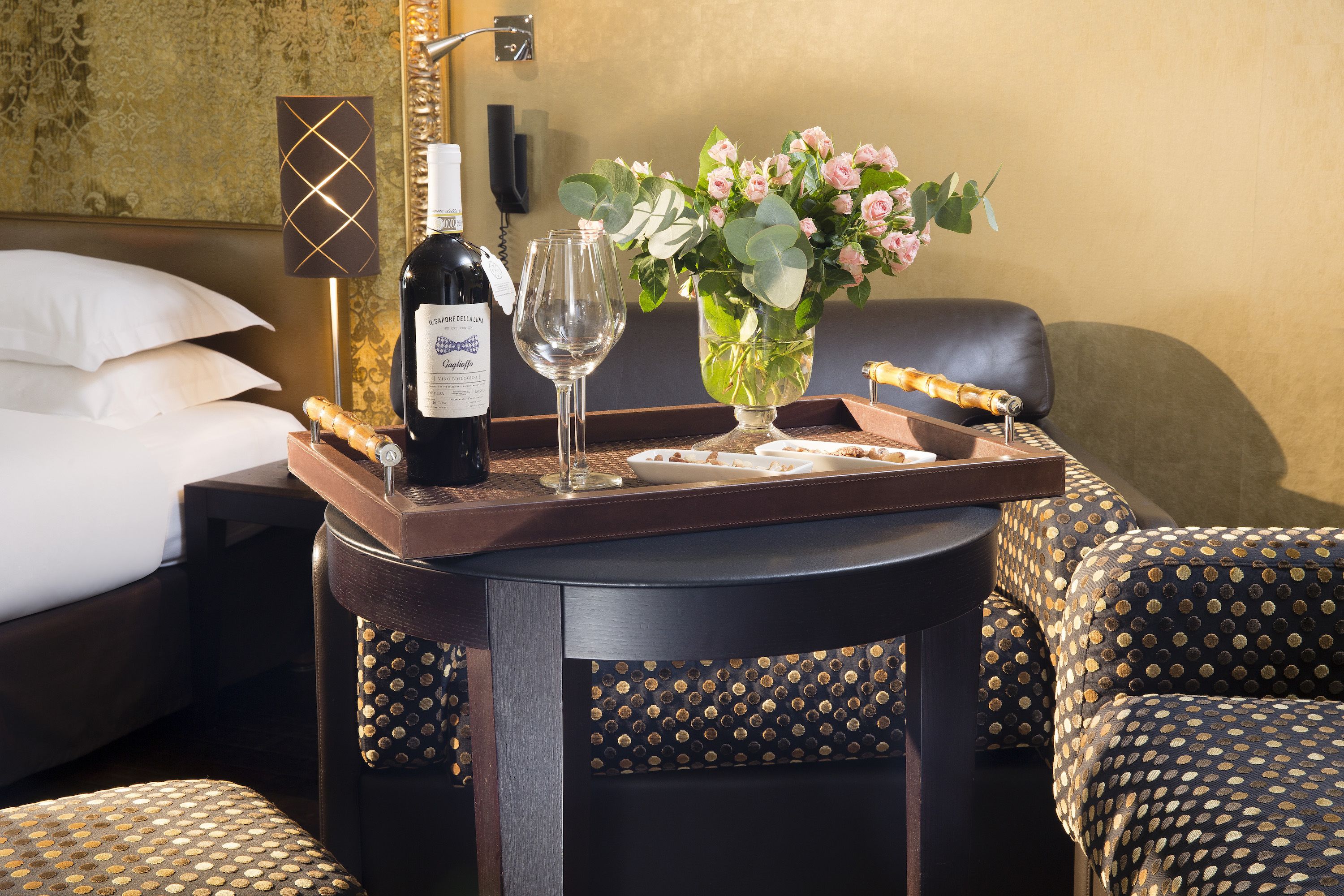 A hotel room with a bed, a bedside table with a bottle of wine, two glasses, a vase with flowers, and a tray with a pastry.