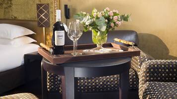 A hotel room with a bed, a bedside table with a bottle of wine, two glasses, a vase with flowers, and a tray with a pastry.