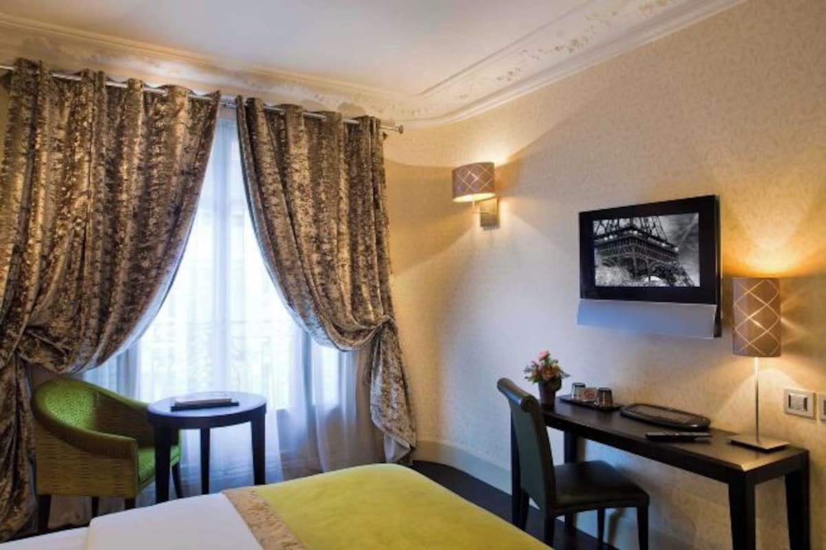 A hotel room with a bed, a desk, two chairs, a small table, curtains, a wall-mounted TV, and a wall-mounted light fixture.