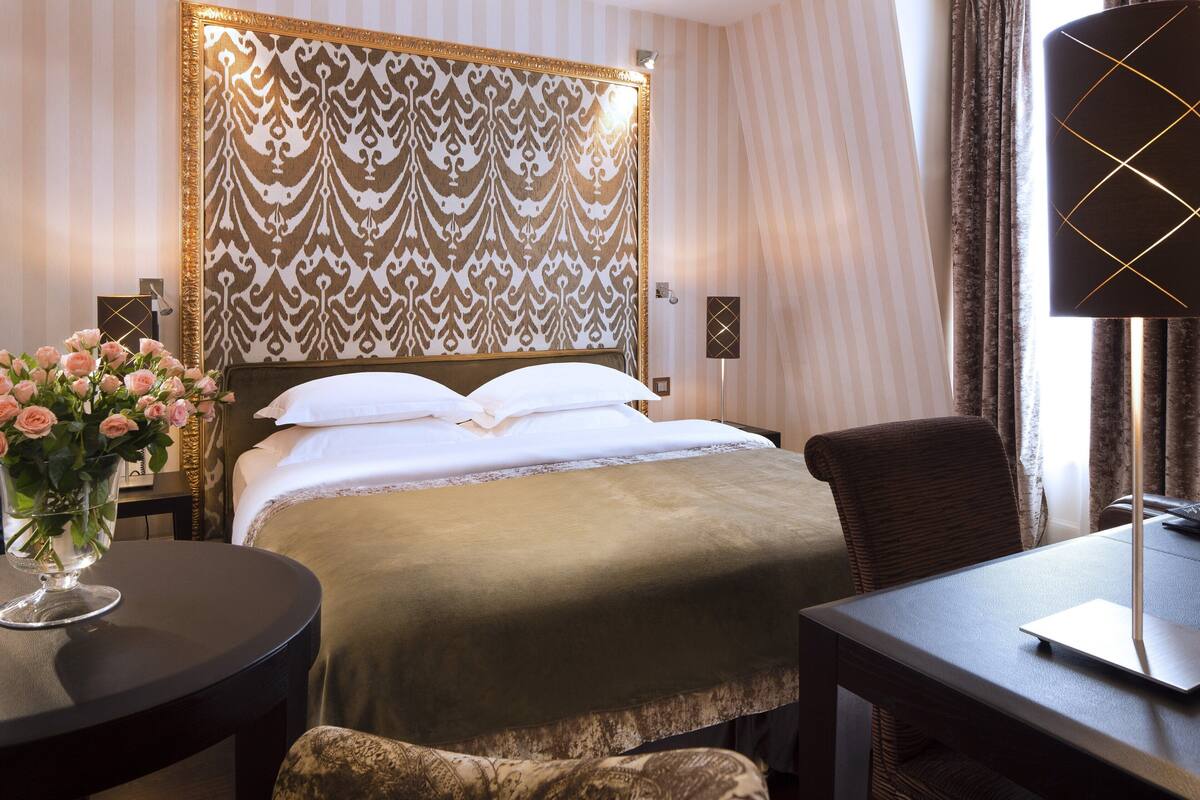 A neatly made bed with white linens, a bedside table with a vase of pink flowers, and a chair at a small table.