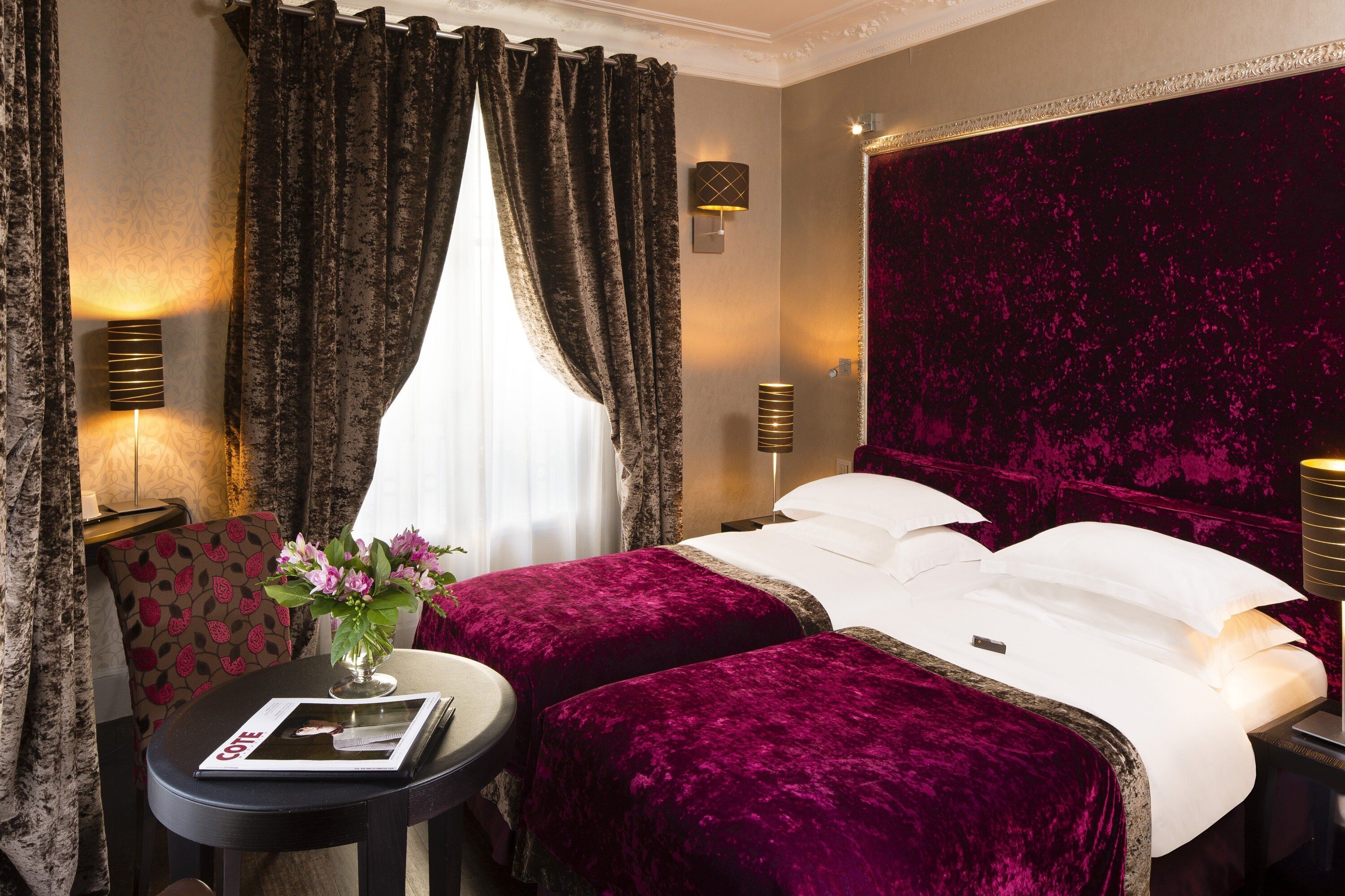 A hotel room with two beds, a sitting area with a table and chair, and a large window with curtains.
