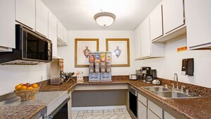 A compact kitchen with white cabinets, a microwave, a coffee maker, and a sink. There are framed pictures on the wall and a basket of oranges on the counter. - Howard Johnson by Wyndham Torrance (Torrance)