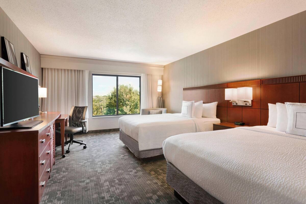 A hotel room with two beds, a desk with a computer, a chair, and a large window with curtains.