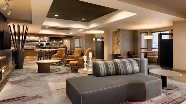 A modern hotel lobby with a sofa, chairs, a coffee table, and a kitchen area in the background. - Courtyard Bloomington Normal (Normal)