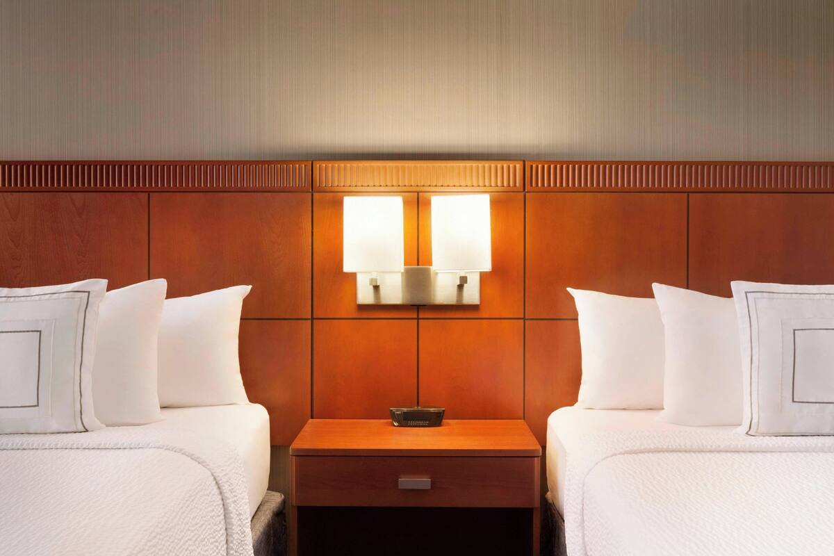 A hotel room with two beds, a nightstand with a lamp, and a wall-mounted light fixture.