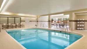 An indoor swimming pool with clear blue water, surrounded by a tiled floor and walls. There are chairs and a table with a view outside. - Courtyard Bloomington Normal (Normal)