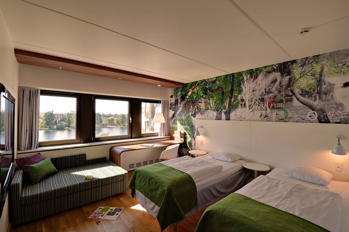 A modern hotel room with a large window, a bed, a sofa, and a mural on the wall.