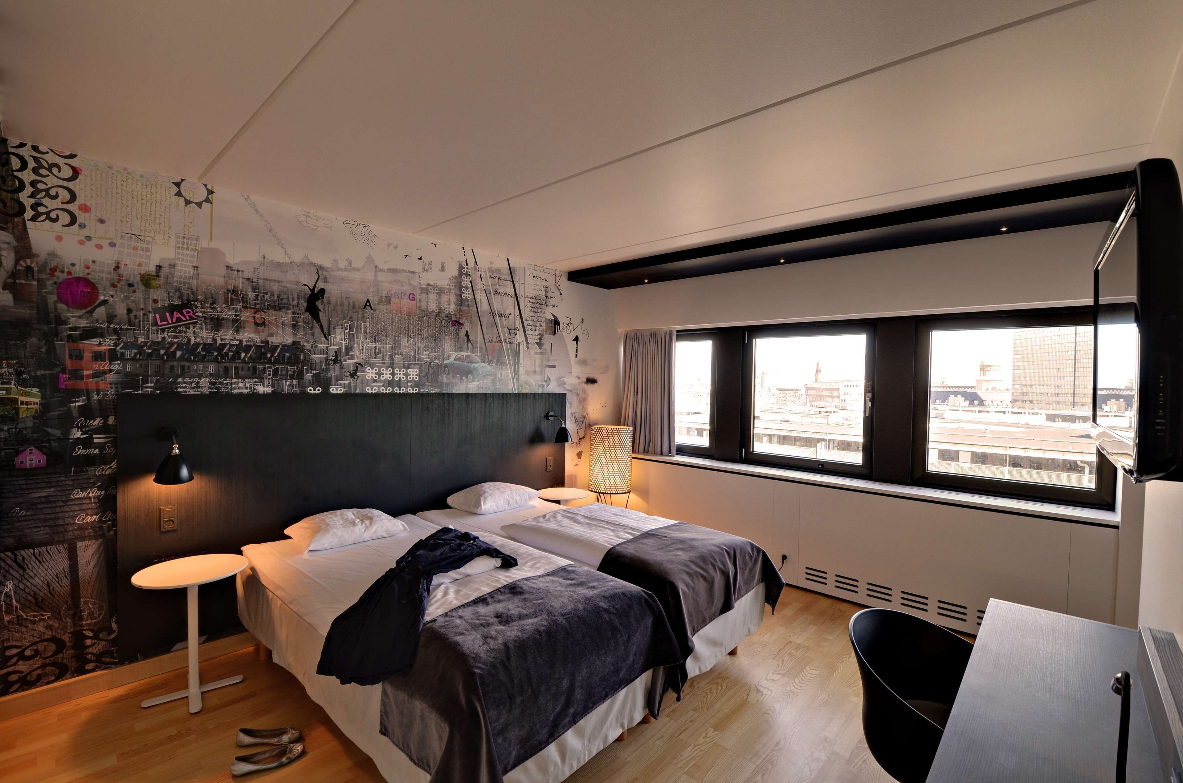 Habitació d'hotel moderna amb un llit gran, una taula de treball, una cadira, una finestra amb vista a la ciutat i una paret amb art abstracte i text.