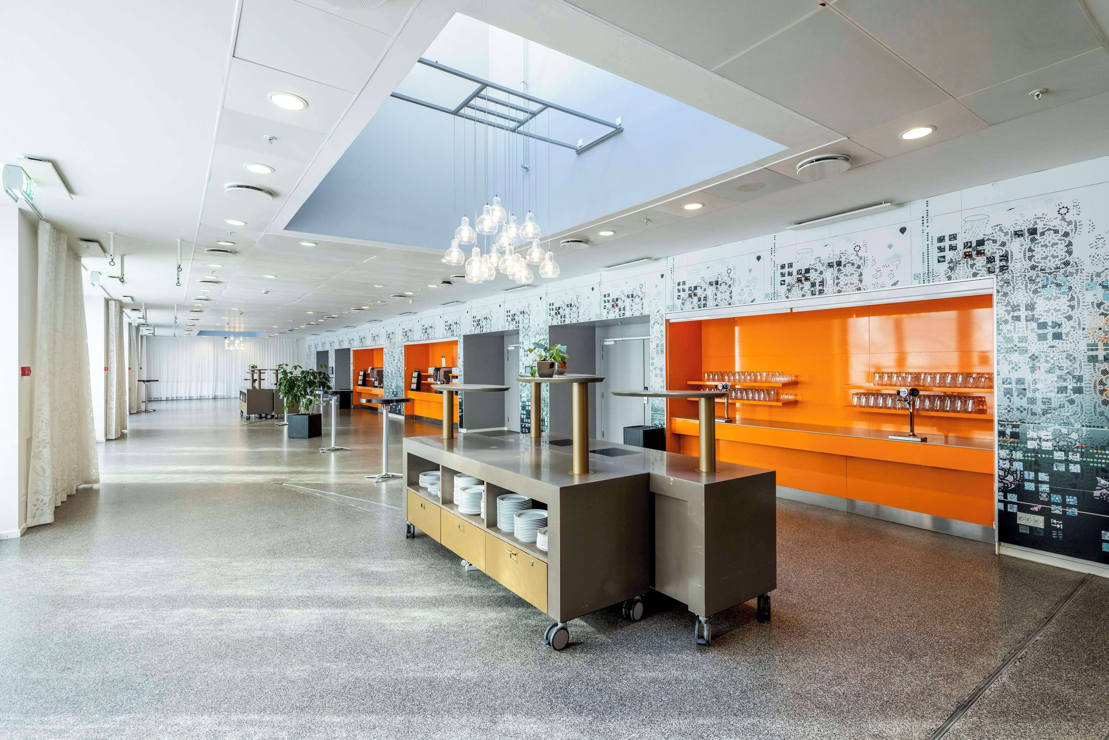 Een ruime lobby met een hoge plafond, moderne verlichtingsarmaturen en een serveerwagen in het midden.