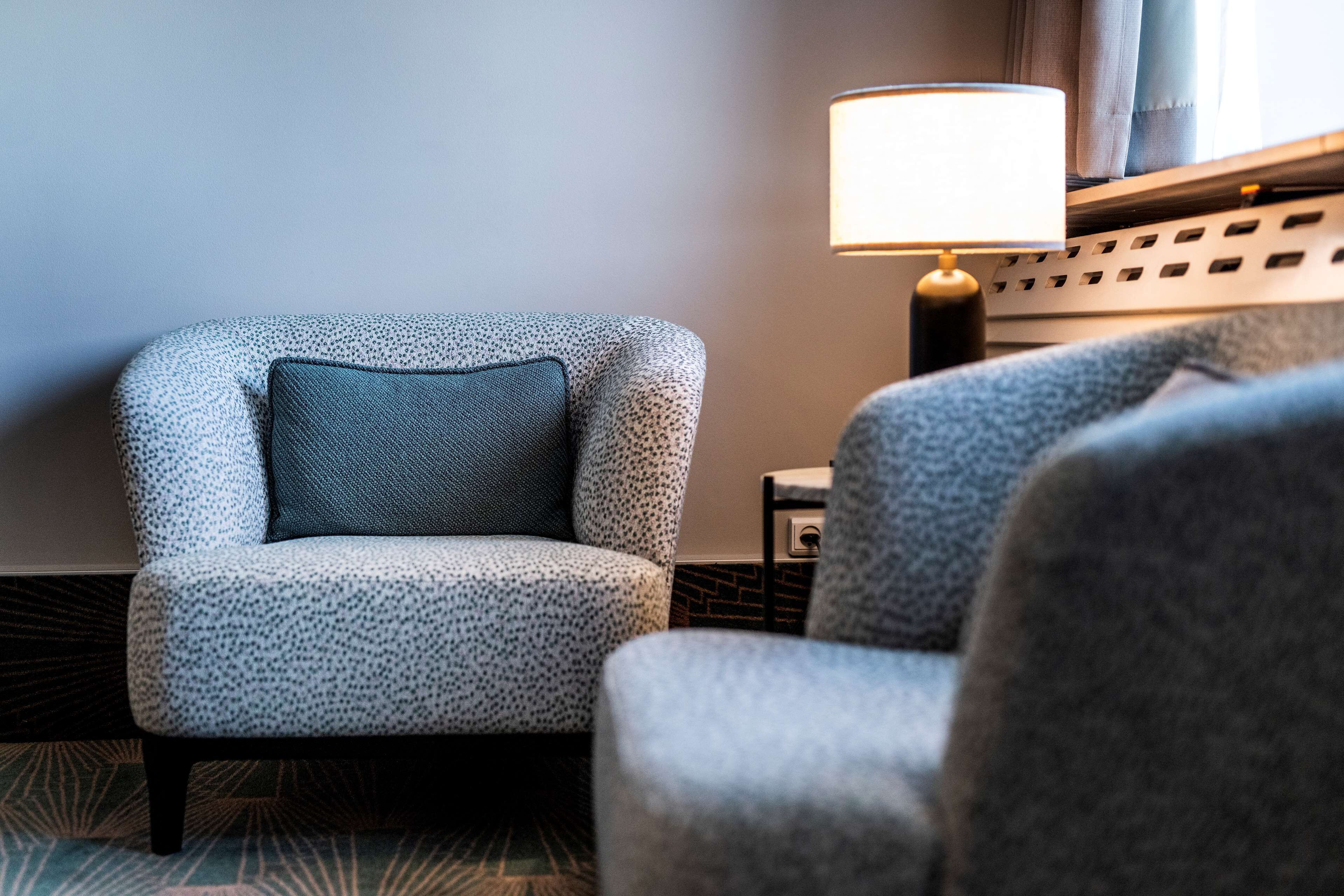 Un sillón moderno con tela estampada y cojín oscuro, situado junto a una lámpara con pantalla blanca.