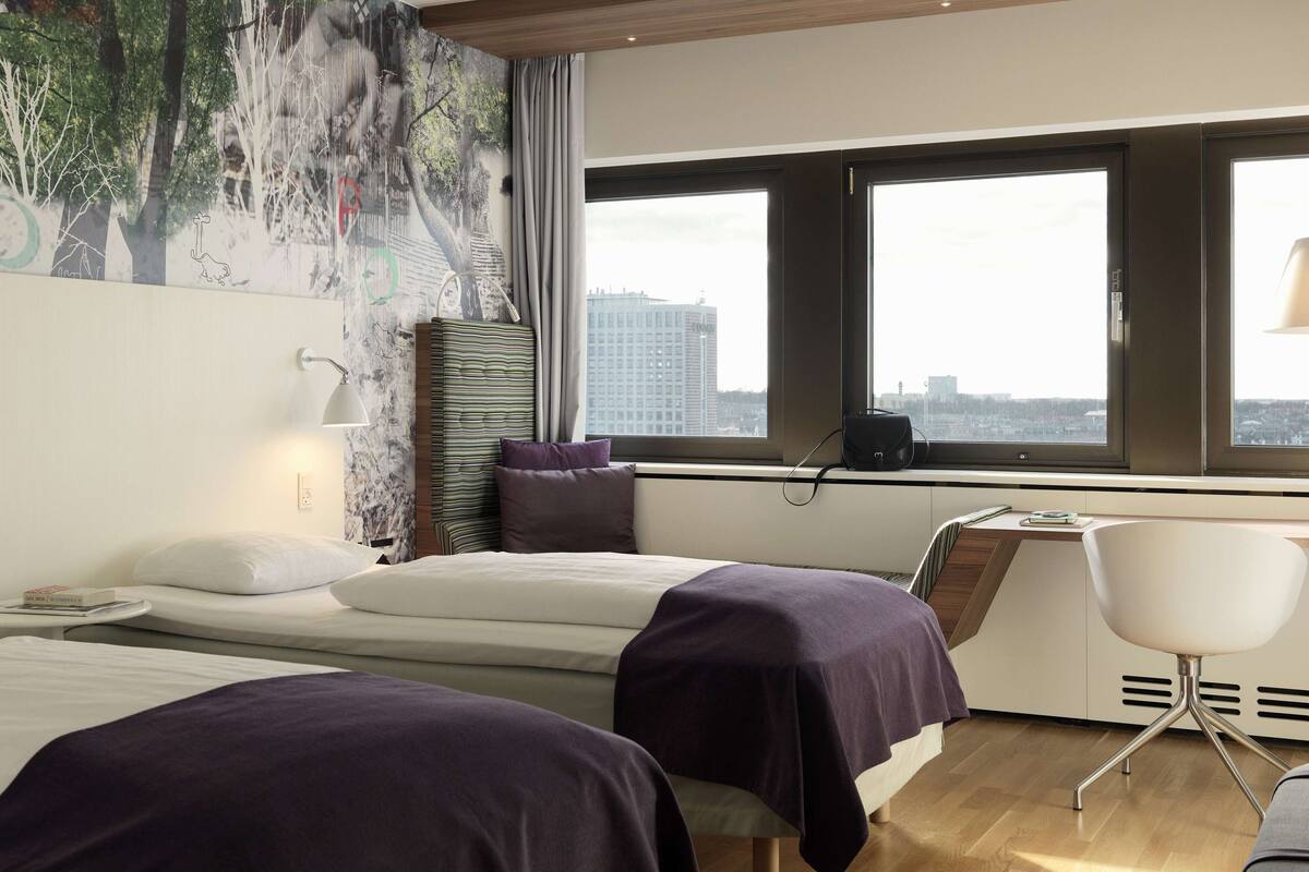 A hotel room with two beds, a desk, a chair, a large window with a city view, and a mural on the wall.