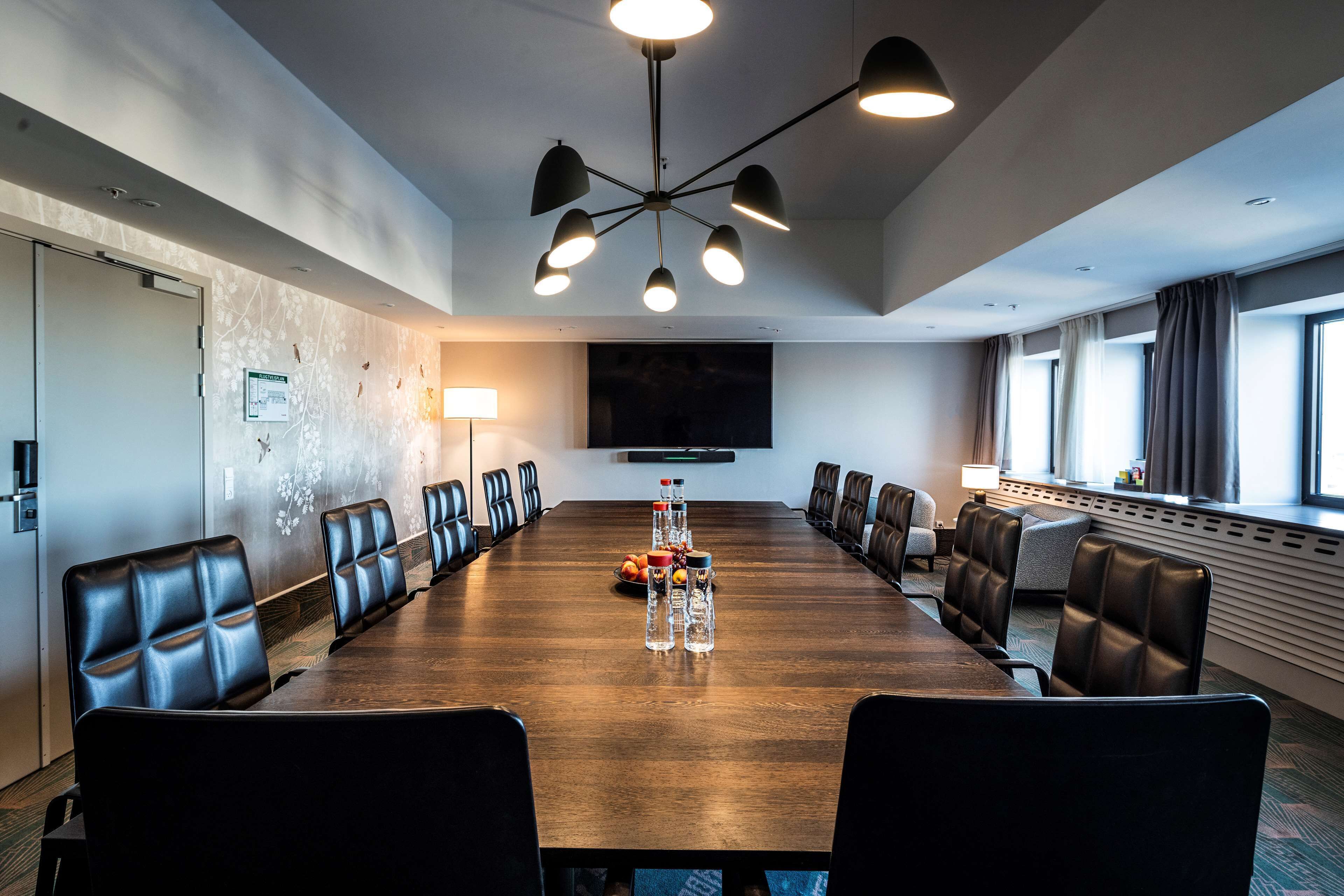A conference room with a long wooden table, black leather chairs, a modern chandelier, and a large flat-screen TV on the wall.
