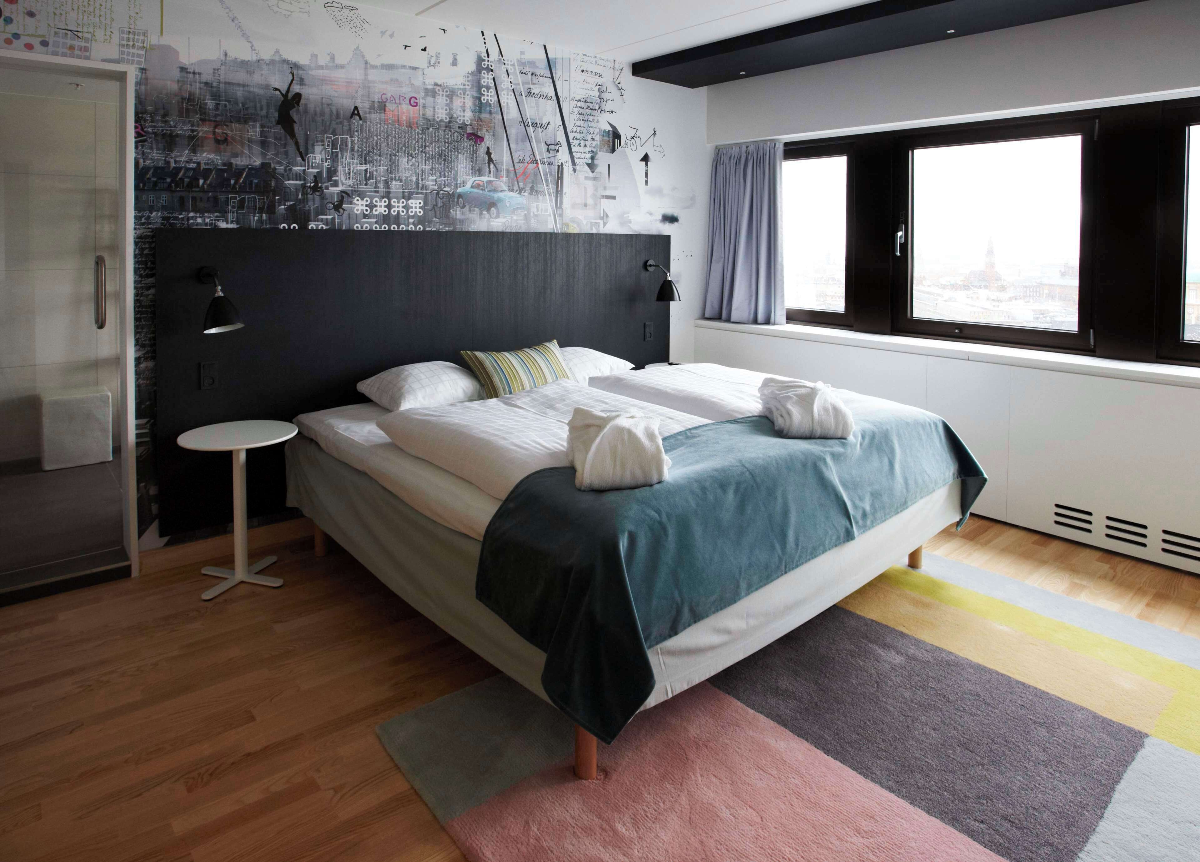 A modern hotel room with a large bed, a small round table, a window with curtains, and a cityscape mural on the wall.