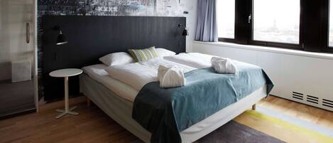 A modern hotel room with a large bed, a small round table, a window with curtains, and a cityscape mural on the wall.