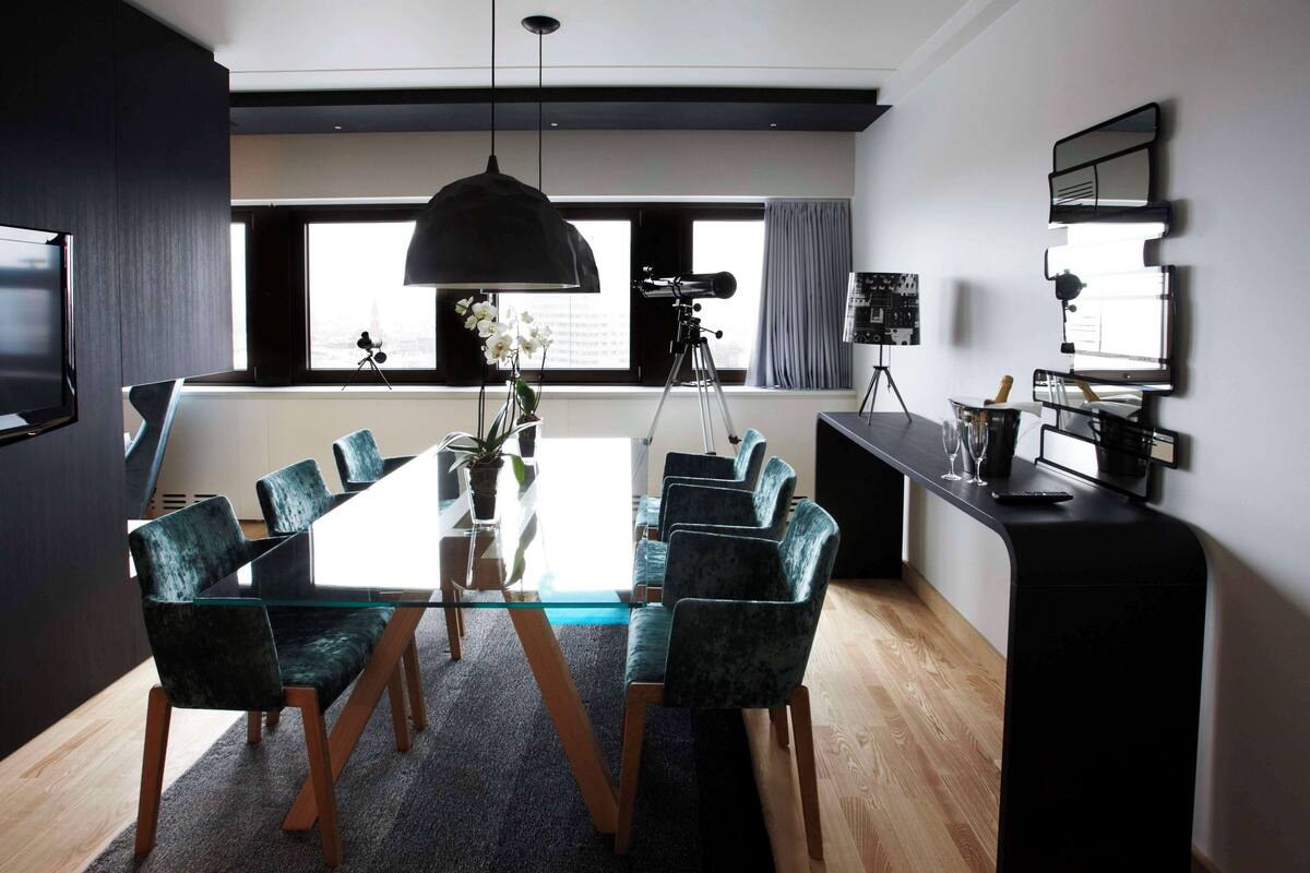 A modern dining area with a glass table, dark upholstered chairs, a black pendant light, and a tripod-mounted camera on a tripod.