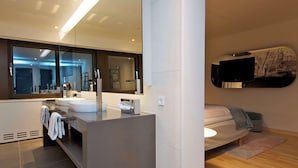 A modern bathroom with a large mirror, two sinks, and a toilet. There is a towel rack with towels, a tissue dispenser, and a wall-mounted heater. The bathroom has a wooden floor and a large window. - Scandic Copenhagen (Copenhagen)