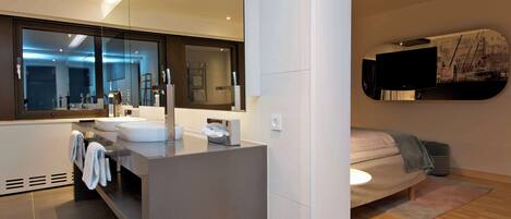 Un baño moderno con un espejo grande, dos lavamanos y un inodoro. Se observa un toallero con toallas, un dispensador de pañuelos y un calentador de pared. El baño tiene piso de madera y un ventanal grande.