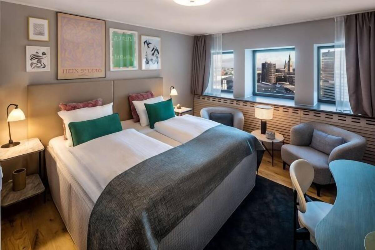 A hotel room with two beds, a sofa, a chair, a small table, and a view of a cityscape.
