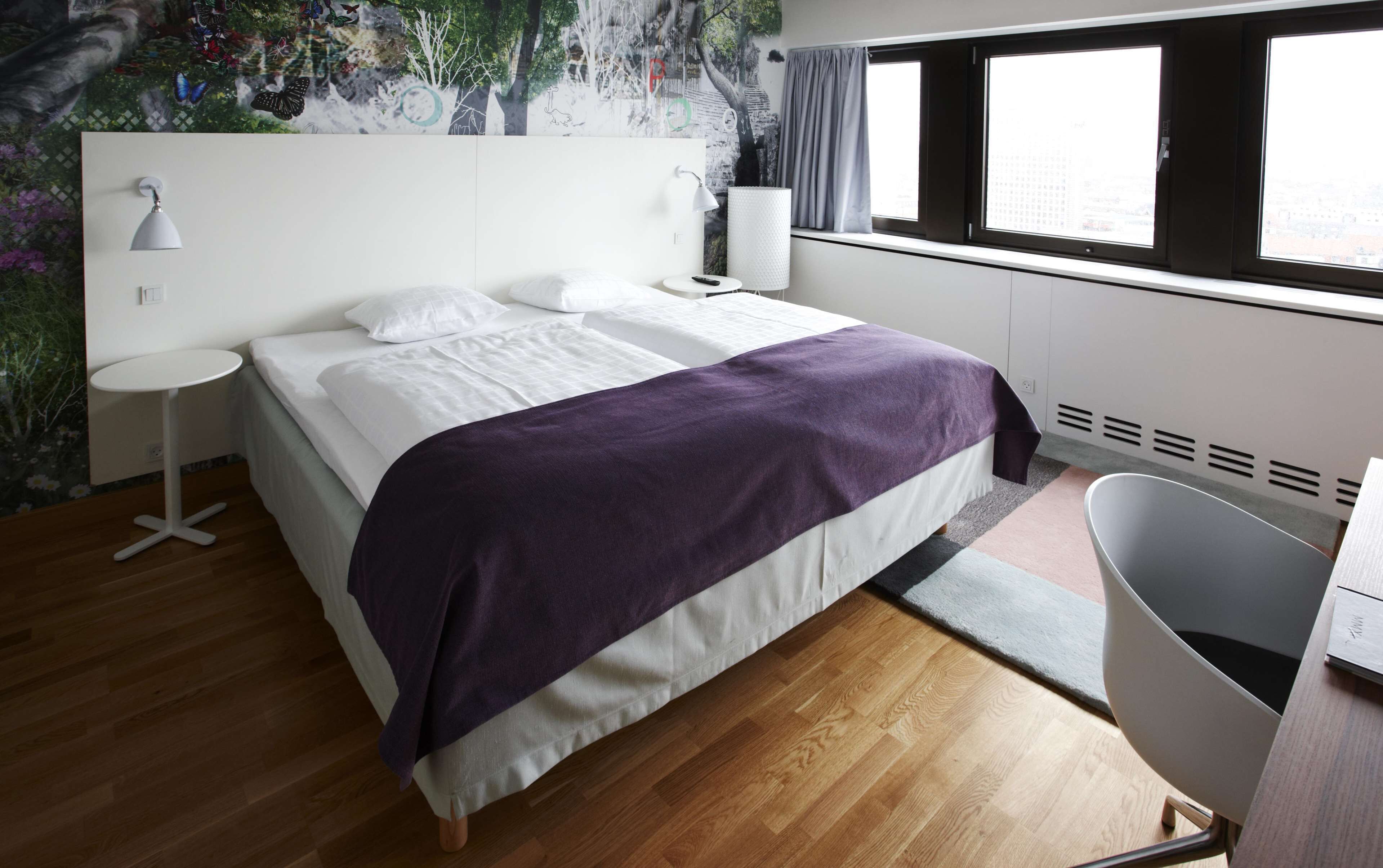 A modern bedroom with a bed, a small round table, a chair, and a large window with curtains.