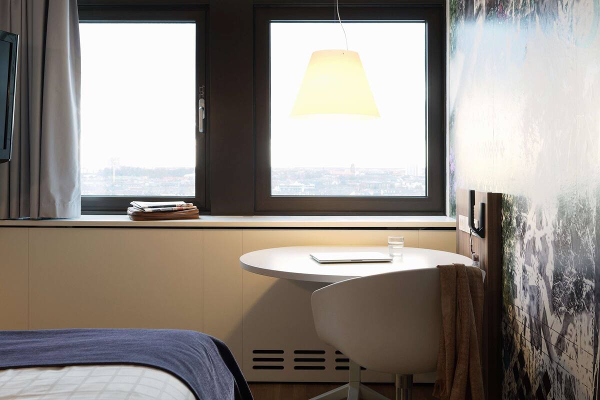 A hotel room with a bed, a round table, a chair, a window with a view of the city, and a hanging lamp.