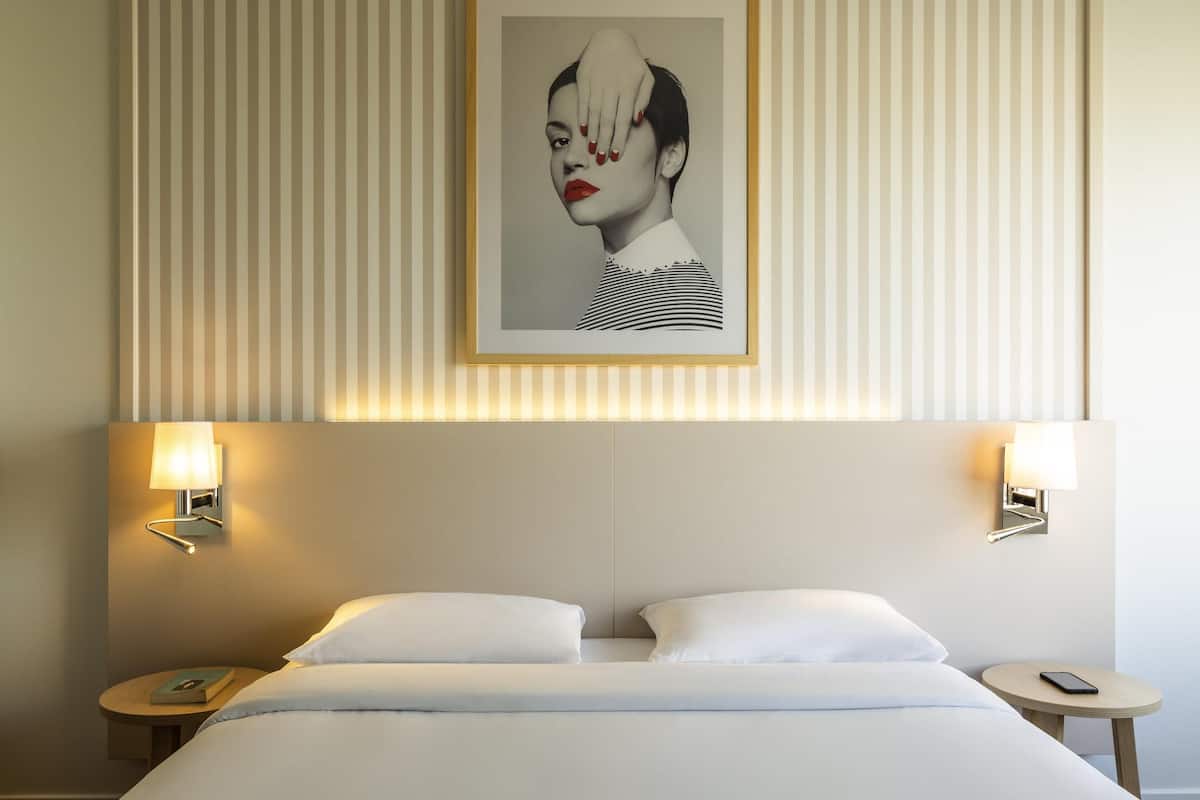 Una habitación de hotel con una cama grande, dos mesitas de noche, una obra de arte enmarcada sobre la cama y lámparas fijadas a la pared.