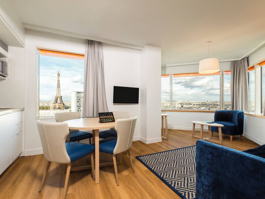 Una habitación de hotel moderna con zona de comedor, sofá, televisión y vistas a la Torre Eiffel.