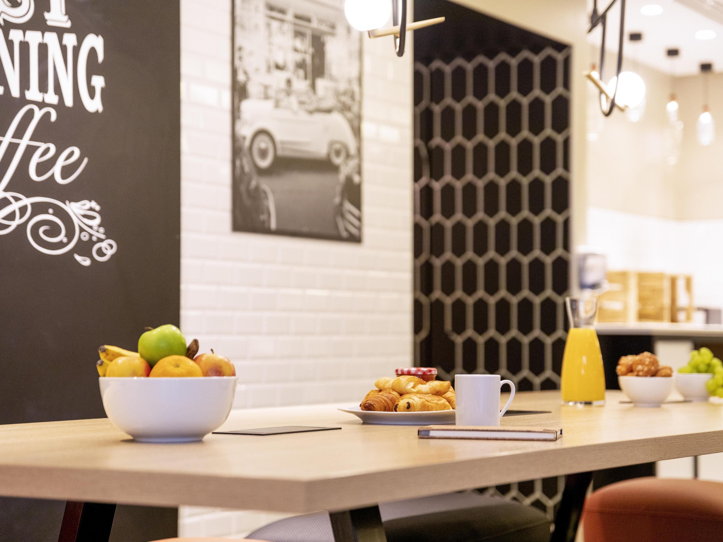 A breakfast table with a bowl of fruit, croissants, a coffee cup, and a glass of orange juice.